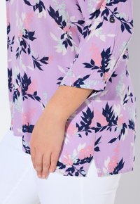 Blusa viola chiaro con motivo floreale in rosa e blu navy, dotata di maniche a tre quarti e spacchi laterali. Tessuto morbido, vestibilità rilassata.