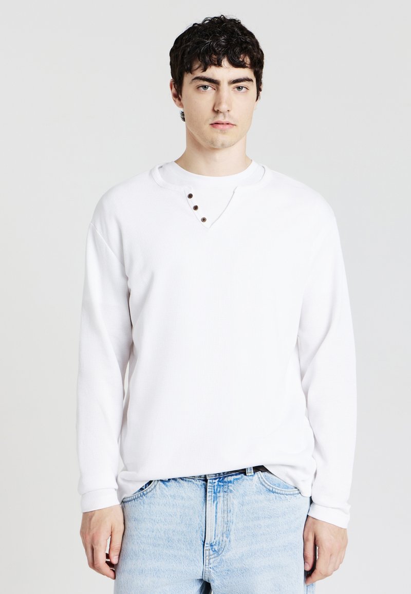 Camicia bianca a maniche lunghe con scollo rotondo e tre bottoni decorativi al colletto. Tessuto intrecciato, abbinata a jeans denim azzurri.