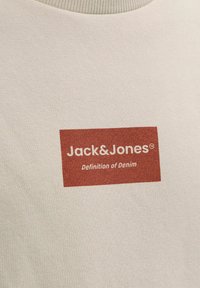 Tissu beige avec un patch logo rectangulaire de couleur rouille portant l'inscription « Jack&Jones Definition of Denim » sur un vêtement à col rond côtelé.