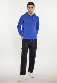 Kék kapucnis pulóver húzózsinórral, fekete cargo nadrággal, fehér varrási részletekkel párosítva. A modellen fehér sportcipők vannak.
