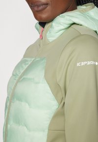 Casaco leve com uma concha verde, apresentando uma parte frontal almofadada em verde menta, fecho éclair rosa em contraste e detalhe do logotipo na manga.