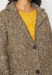 Hn�ědý a krémový kabát s výrazným vzorem, límcem a černými knoflíky, nošený přes žlutý košilový top. Měkká pletená tkanina.