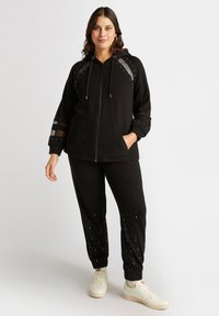 Svart zip-hoodie med texturerade detaljer och rhinestones, parat med matchande joggers som har rhinestones på sidan, och vita sneakers.