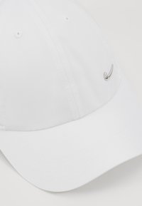 Fehér pamut baseballsapka, ívelt karimával, varrott részletekkel és egy kis fém logóval az elején.
