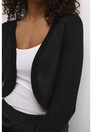 Cardigan noir court en tissu texturé, à l'avant ouvert et avec des manches trois-quarts, superposé à une chemise blanche.