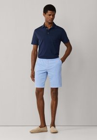 Homme portant un polo à manches courtes bleu marine, un short bleu clair et des chaussures beige sans lacets, debout avec une main dans la poche.