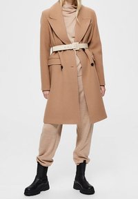 Dlouhý velbloudí kabát s páskem v pase, s dvojitým zapínáním a velkými klopami. Kombinovaný s béžovým rolákem a černými kotníkovými botami.