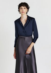 Donna con capelli corto e ricci, che indossa una camicia satinata blu navy con bottoni e una gonna satinata alta in vita di colore grigio scuro, con anelli d'argento sulla mano.