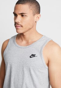 Pelēka bezpiedurkņu bluze, izgatavota no kokvilnas maisījuma, ar apaļu kakla izgriezumu un melnu Nike logo kreisajā krūtī. Gluda tekstūra.