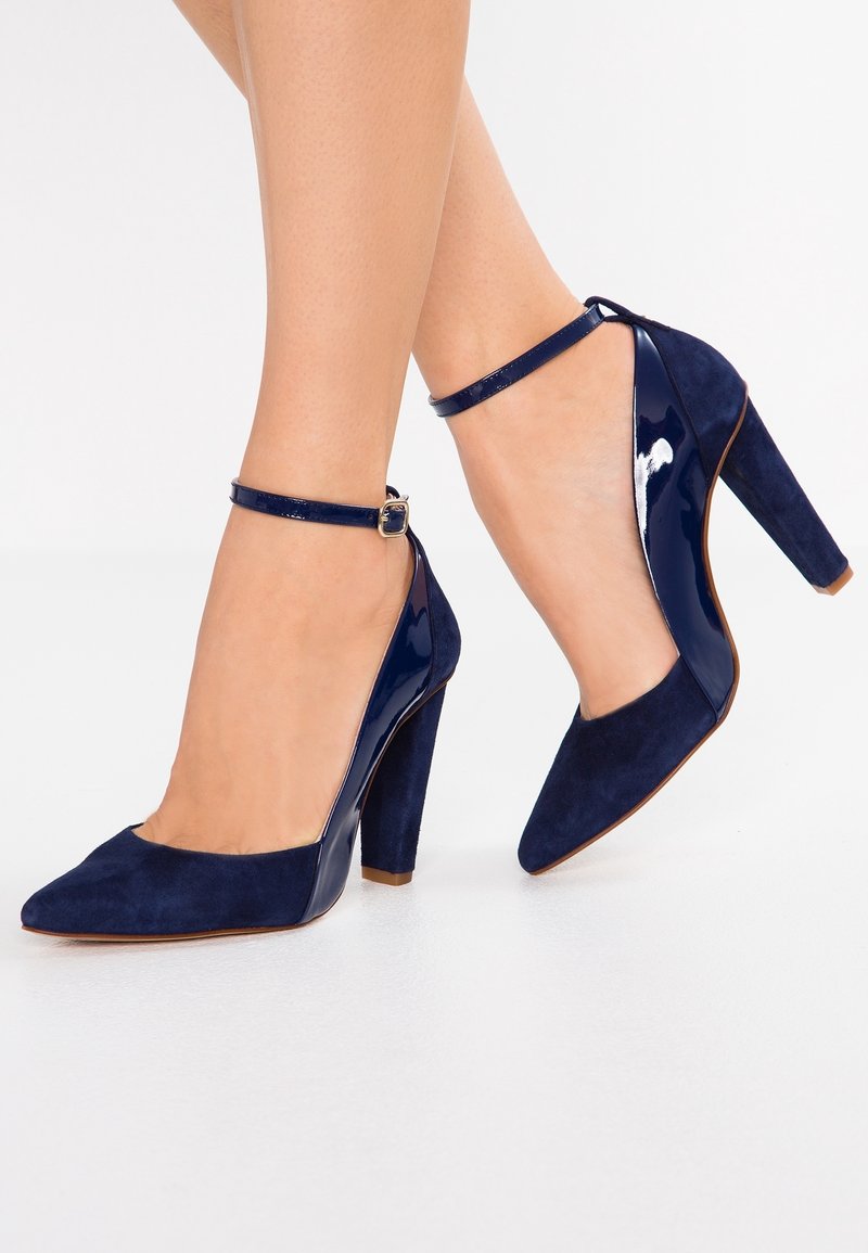 Deux jambes portant des chaussures à talons hauts bleu marine à bouts pointus, avec des brides à la cheville et un mélange de textures en suède et cuir verni.