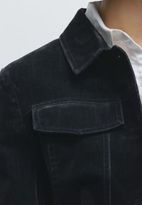 Giacca in denim nero con colletto appuntito e taschino sul petto. Il tessuto presenta una trama sottile con dettagli di cuciture azzurro chiaro.