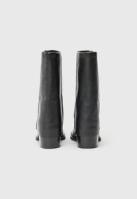 Un par de botas de cuero negras hasta el tobillo con tacones cuadrados, vistas desde atrás sobre un fondo gris claro liso.