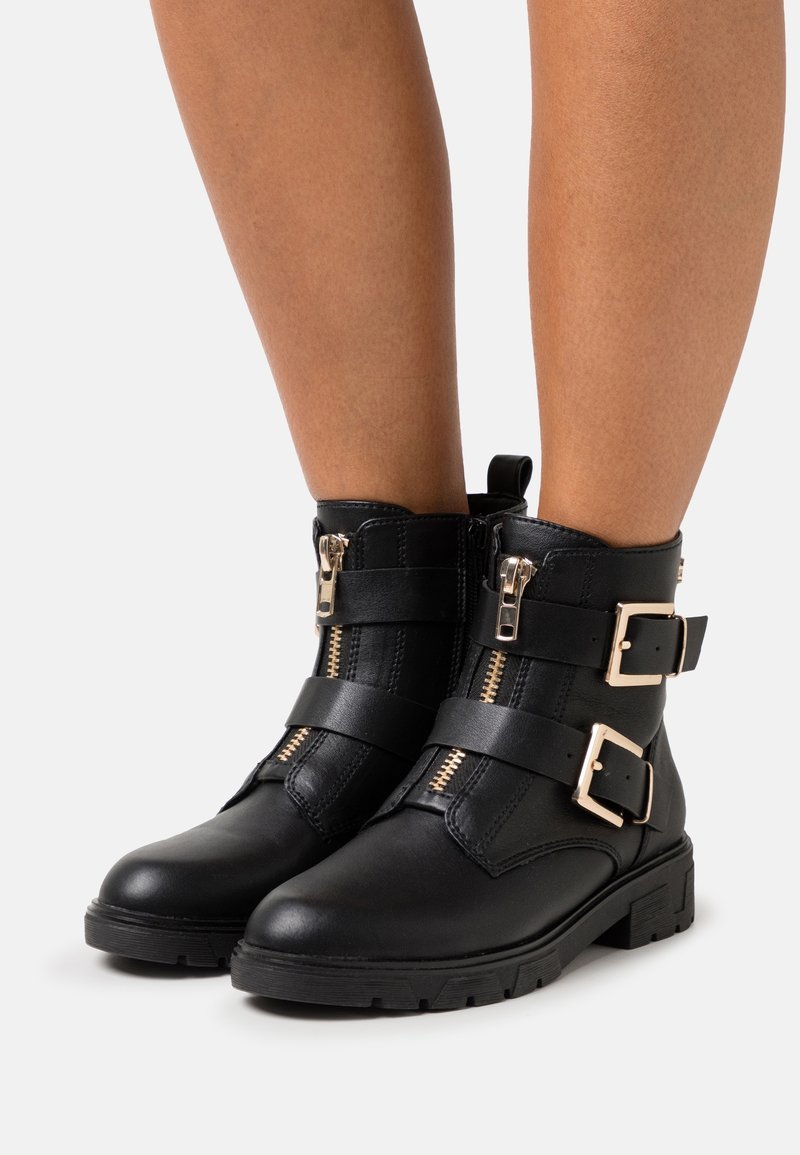 Bottines noires avec double boucle dorée et fermetures éclair à l'avant, portées sur des jambes nues, présentées sur un fond clair uni.