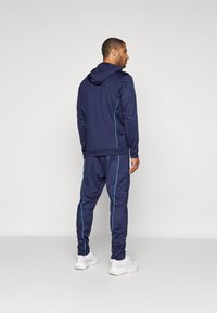 Marinebukse med kontrasterende lyseblå sømmer, med en hettejakke og tapered bukser, laget av glatt og elastisk stoff. Hvite joggesko.
