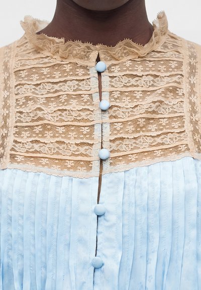 Άτομο με σκούρο δέρμα που φοράει ένα γαλάζιο φόρεμα με πλισέ φούστα και μπεζ δαντελένιο πάνω μέρος, κλειστό με κουμπιά καλυμμένα με γαλάζιο ύφασμα.