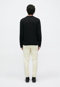 Görögödő sötét hajú férfi, fekete hosszú ujjú inget, bézs jogger nadrágot és fekete cipőt viselve, háttal egy egyszínű fehér háttér előtt.