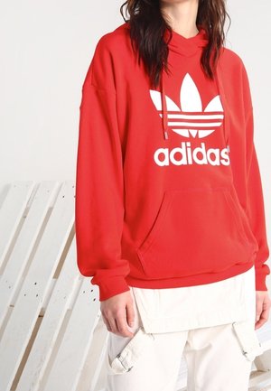 Osoba ubrana w czerwoną bluzę z kapturem Adidas z białym logo i białe spodnie, stojąca przy białym drewnianym tle palety.