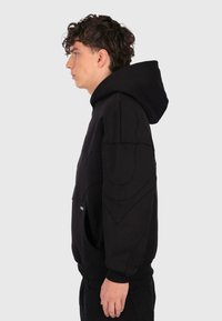 Junger Mann mit lockigem Haar, der einen schwarzen Hoodie und schwarze Hosen trägt, im Profil vor einem schlichten hellen Hintergrund stehend.