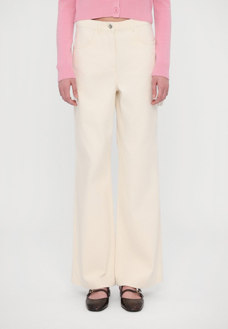 Wide-leg, cream-colored pants made from cotton blend, featuring front pockets and a button closure. Paired with brown flat shoes.