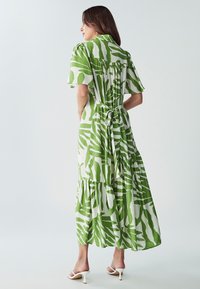 Maxi šaty v bílé barvě s zelenými listovými vzory, s krátkým rukávem, s páskem na zavázání a s vrstvenými volány na lemu. Hladká textura látky.