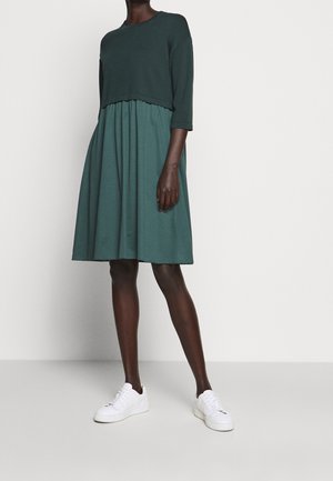 Femme debout portant un haut à manches longues vert foncé sur une robe-jupe bleu sarcelle mi-longue et des baskets blanches sur un fond uni.