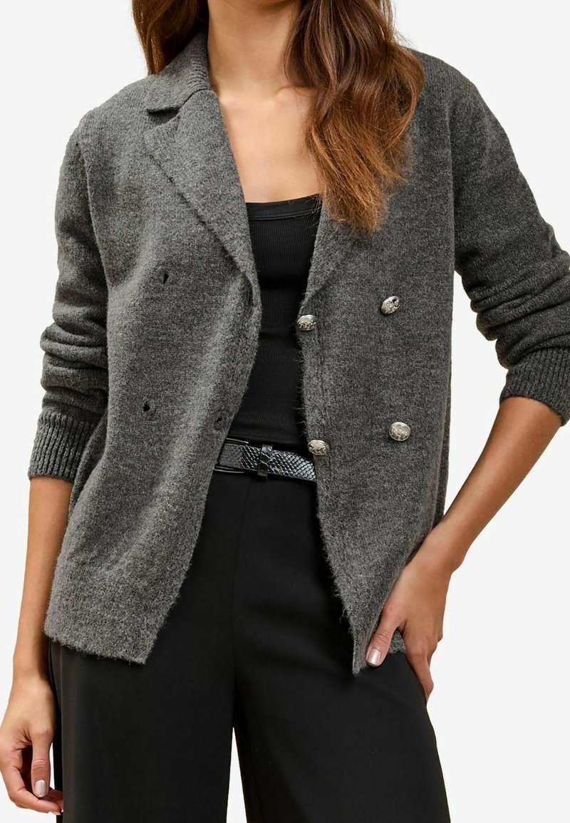 Femme portant un cardigan en maille gris à double boutonnage sur un top noir sans manches et un pantalon noir avec une ceinture texturée.