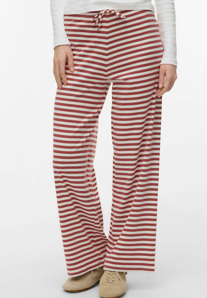 Personne portant un pantalon ample à rayures horizontales rouges et blanches avec une taille à cordon de serrage et des chaussures beiges à lacets, debout devant un fond uni.