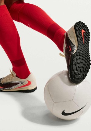 Spiller iført røde sokker og beige Nike fodboldstøvler balancerer en hvid Nike fodbold med en fod mod en hvid baggrund.