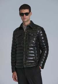 Schwarze gepolsterte Jacke mit glänzender Oberfläche, Druckknopfverschluss, zwei Brusttaschen und Punktkragen. Getragen über einem leichten Hemd und einer Hose.