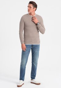 Camicia a maniche lunghe di un beige chiaro con una texture, con colletto bianco, abbinata a jeans blu e sneaker bianche con dettagli marroni.