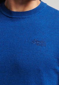 Pull bleu tricoté avec un col ras du cou côtelé. Présente un subtil logo "SuperDry" brodé sur la poitrine. Texture douce.