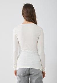 Langärmliges, elfenbeinfarbenes Oberteil aus leichtem Stoff, mit weitem Rundhalsausschnitt und figurnahen Schnitt, kombiniert mit grauen Jeans.