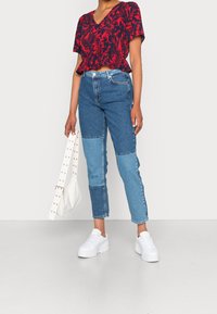 Rotes Blumenmuster Kurzarmoberteil, blaue Patchwork-Jeans, weiße Sneakers und eine weiße Umhängetasche mit metallischen Akzenten.