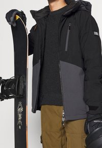 Doudoune noire et grise avec une fermeture éclair, pull texturé foncé, pantalon cargo olive, et snowboard avec des motifs graphiques et un bord orange.