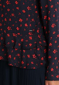 Blusa floreale in tessuto blu navy con motivi a fiori rossi, caratterizzata da un design a strati e un'etichetta del marchio discreta all'orlo. Maniche lunghe, texture leggera.