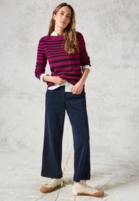 Maglione a righe in bordeaux e rosa scuro, indossato sopra una camicia bianca con colletto. Abbinato a pantaloni a gamba larga blu navy e sneakers beige.