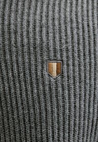 Ribbed gray fabric with a textured appearance. Features a small embroidered shield badge in brown and beige at the bottom corner.