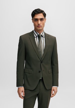 Traje verde oliva con dos botones, solapas tipo muesca y bolsillos laterales. Acompañado de una camisa de rayas gruesas y una corbata texturizada en tonos a juego.
