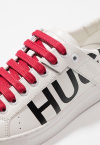 Baskets en cuir blanc avec un audacieux logo "HUGO" en noir, des lacets en tissu rouge et des œillets ronds. Texture lisse avec des accents de design superposés.
