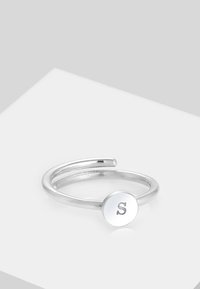Ezüst gyűrű nyitott designnal, sima kör alakú tárcsával, amelyen az "S" betű van gravírozva. Fényes felület, minimalista stílus.