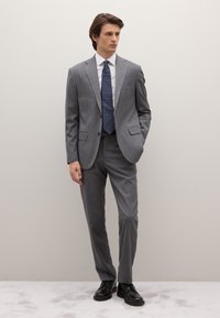 Abito grigio in tessuto strutturato, composto da blazer a due bottoni, pantaloni sartoriali, camicia bianca e cravatta scura a pois. Scarpe nere.