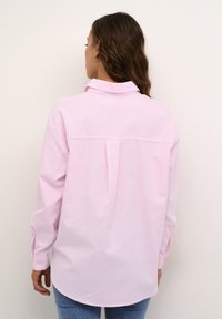 Chemise à manches longues en coton rose clair. Dotée d'un col, d'un pli dans le dos et d'un ourlet arrondi. Associée à un jean bleu.