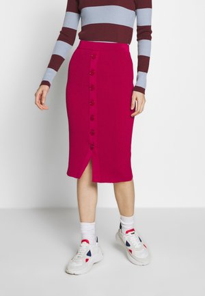 Femme portant une jupe magenta côtelée longueur genou avec des boutons sur le devant, associée à un haut à manches longues rayé et des baskets blanches avec des chaussettes.
