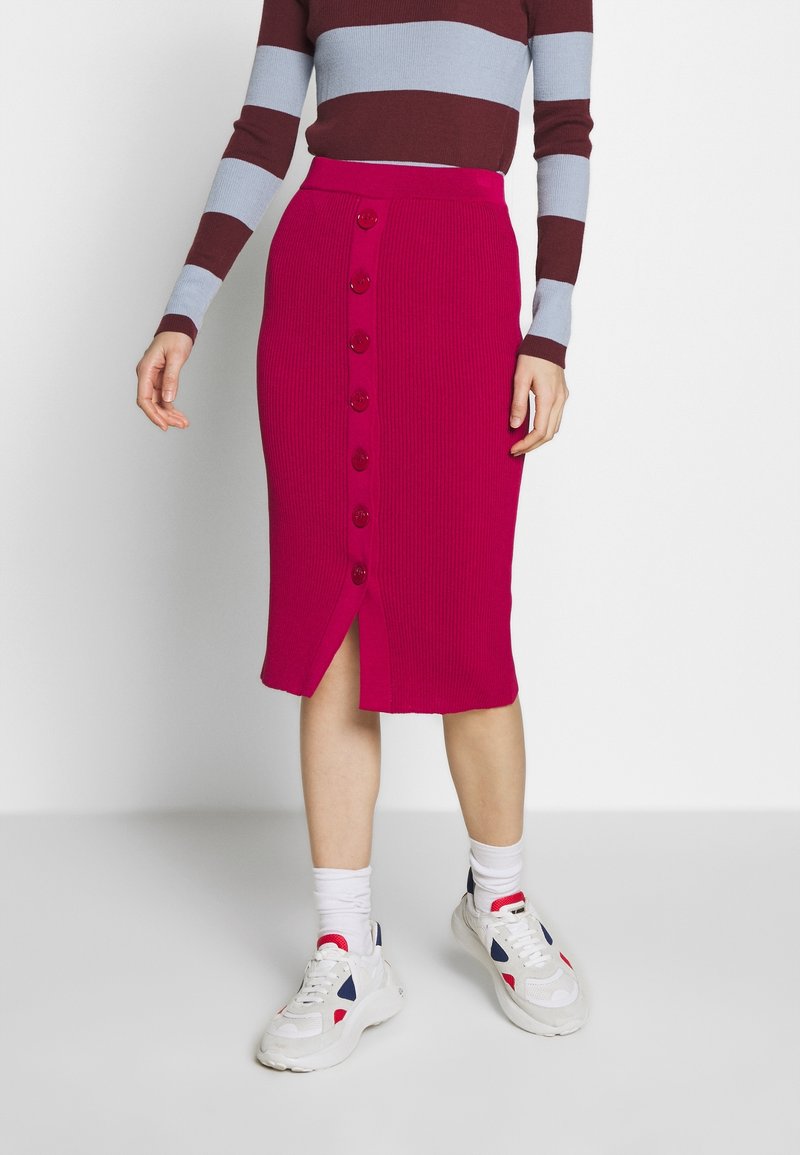 Femme portant une jupe magenta côtelée longueur genou avec des boutons sur le devant, associée à un haut à manches longues rayé et des baskets blanches avec des chaussettes.