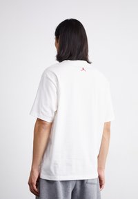 T-shirt bianco in cotone con maniche corte e scollo rotondo, con un piccolo logo rosso sulla parte superiore della schiena. Tessuto morbido e vestibilità rilassata.