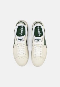 Beige leather sneakers with green accents, white laces, and perforated toe. Features logo on tongue and textured fabric interior.