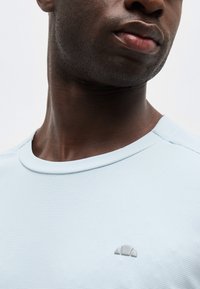 Κοντινό πλάνο ενός ατόμου που φοράει ένα ανοιχτό μπλε πουκάμισο με υφή, με ένα μικρό ημικυκλικό λογότυπο στο στήθος, δείχνοντας μέρος του λαιμού και του κάτω μέρους του προσώπου.