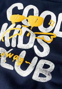 Tumedat sinised kangad, millel on julged, kulunud valged kirjad "COOL KIDS CLUB", kollased prillid ja pintslitõmbe aktsendid.