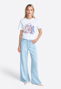 T-shirt blanche avec un graphique de panier coloré ; associé à un jean large bleu clair. La mannequin se tient avec une main sur la hanche.
