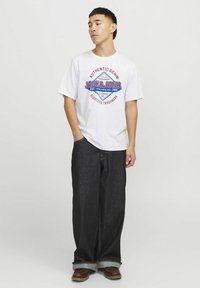 Witte T-shirt met een rood en blauw grafisch ontwerp met de tekst "JACK & JONES." Gecombineerd met losse, donkere denim jeans en bruine schoenen.